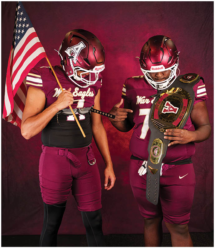 Two people in maroon football uniforms stand against a dark red background, one holding an American flag and the other holding a championship belt. Description generated by AI