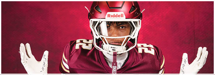 A person wearing a maroon football uniform and helmet with the number 22 raises both hands against a red background. Description generated by AI