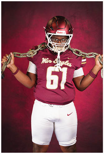 A person wearing a maroon football helmet and jersey with “War Eagles" written on it is holding a heavy chain over their shoulder against a maroon background. Description generated by AI