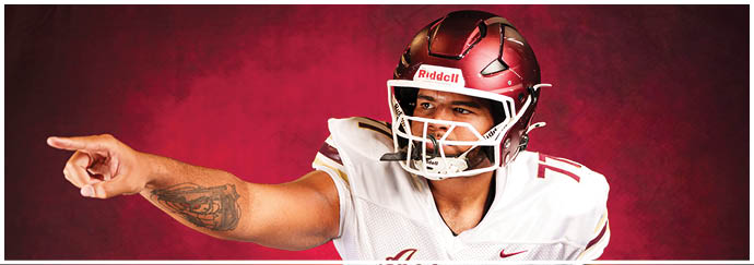A person wearing a white football jersey with the word “Astronaut" and the number 77 is in a crouched position with one arm extended forward against a red background. Description generated by AI