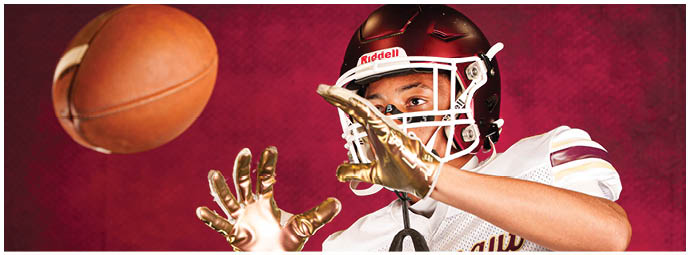 A person wearing a football helmet and gloves is reaching out to catch a football against a red background. Description generated by AI