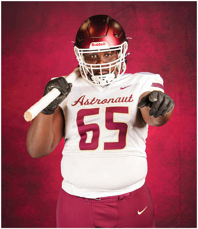 A person wearing a football uniform with the number 65 and the word Astronaut on the jersey is holding a bat over one shoulder and pointing forward against a red background. Description generated by AI