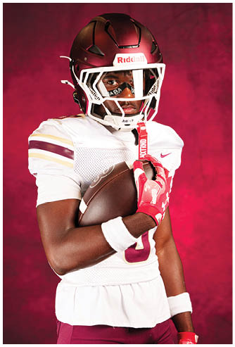 A person wearing a maroon football helmet and uniform holds a football while posing against a red background. Description generated by AI