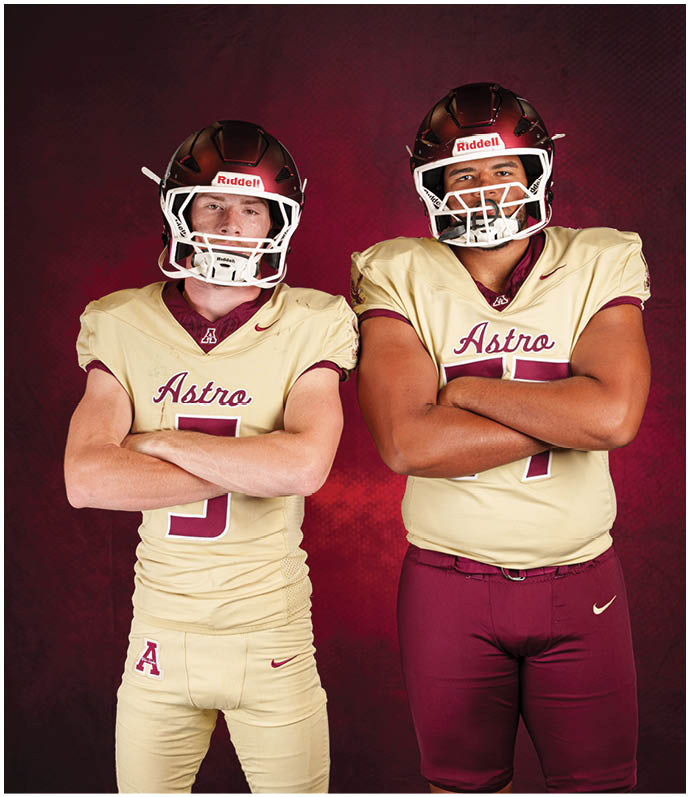 Two people wearing football uniforms with helmets and crossed arms stand against a dark red background. Description generated by AI