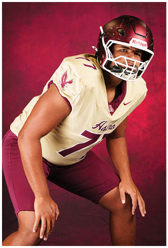 A person wearing a maroon and cream football uniform with a helmet is posed in a crouched stance against a maroon background. Description generated by AI