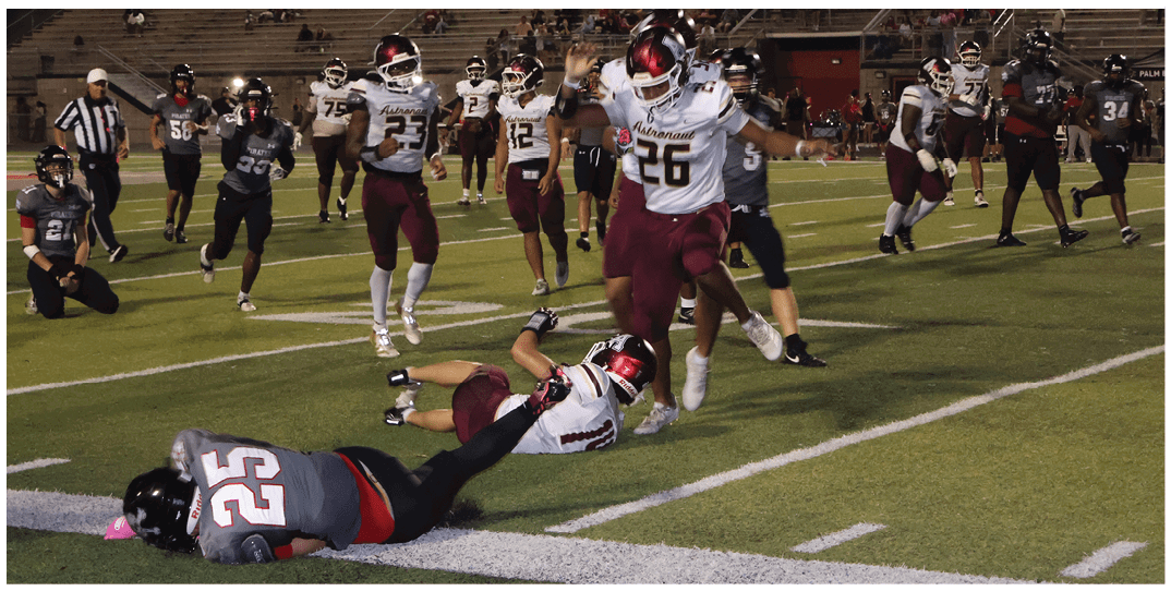 Two people in football uniforms are on the ground near the sideline with one person holding the other's leg while several others stand nearby on a football field. Description generated by AI