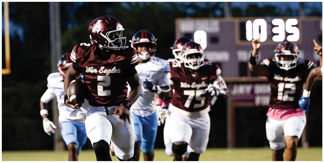 A person wearing a maroon football uniform with the number 2 is running with the ball on a football field while other players in maroon and white uniforms chase behind. Description generated by AI