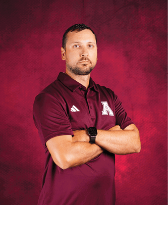 A person with short hair and a beard is standing with arms crossed wearing a maroon polo shirt with a white letter A and an Adidas logo against a maroon textured background. Description generated by AI
