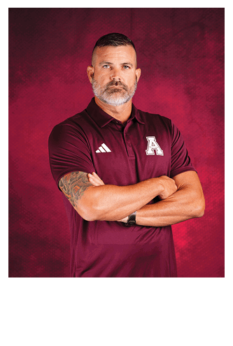 A person with short hair and a beard wearing a maroon polo shirt with an “A" logo and Adidas stripes stands with arms crossed against a red background. Description generated by AI