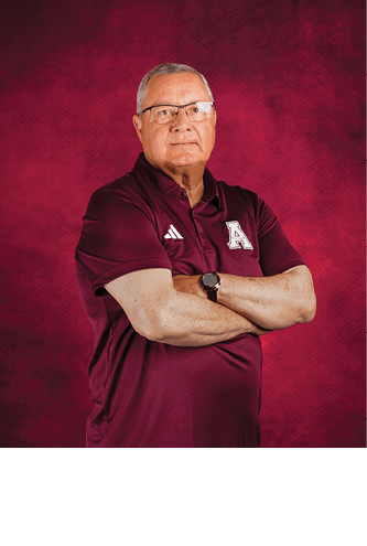 A senior person wearing glasses and a maroon polo shirt with a logo stands with arms crossed against a textured maroon background. Description generated by AI