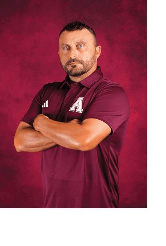 A person with short curly hair and a beard is standing with arms crossed wearing a maroon polo shirt with a white letter A on the chest against a maroon textured background. Description generated by AI