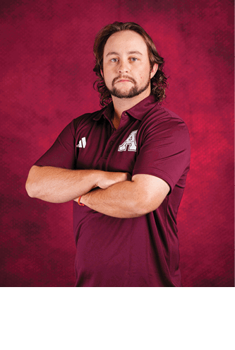 A person with shoulder-length wavy hair and a beard is standing with arms crossed wearing a maroon polo shirt with a white letter A on the chest against a textured maroon background. Description generated by AI