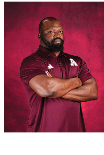 A person with a beard wearing a maroon polo shirt with a white letter A on it stands with arms crossed against a textured red background. Description generated by AI