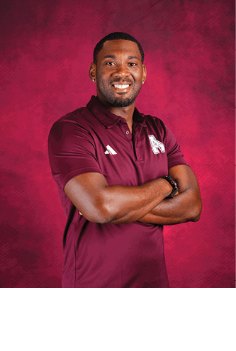 A person wearing a maroon polo shirt with logos stands confidently with arms crossed against a textured maroon background, smiling. Description generated by AI