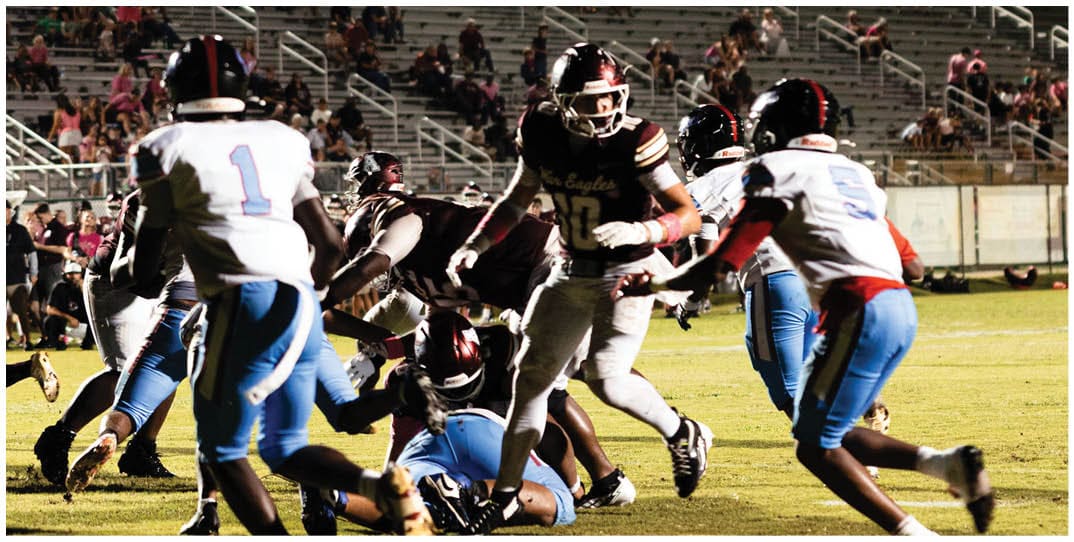 People wearing football helmets and uniforms are engaged in a nighttime football game on a field with bleachers and spectators in the background. Description generated by AI
