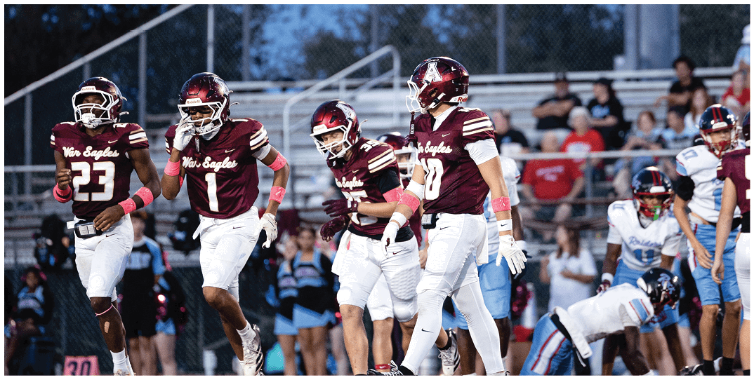 Several people wearing maroon football uniforms with helmets are on a field with a chain-link fence and spectators in the background. Description generated by AI