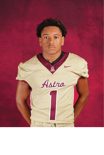 Person with short curly hair wearing a beige football jersey with the word Astro and the number 1 on the front standing against a red textured background. Description generated by AI