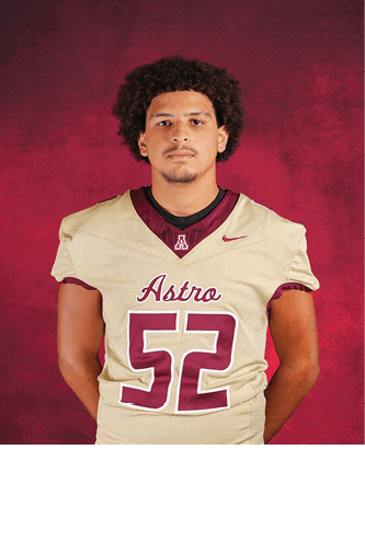 A person with curly hair wearing a beige football jersey with the number 52 and the word Astro stands against a red textured background with hands behind their back. Description generated by AI