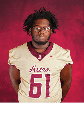 A person with glasses and short dreadlocks is wearing a beige and maroon football jersey with the word Astro and the number 61 on the front, standing against a red textured background. Description generated by AI
