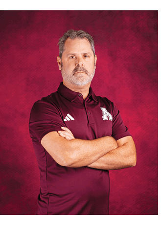 A person with short hair and a beard stands with arms crossed wearing a maroon polo shirt with a white “A" logo and Adidas stripes against a red textured background. Description generated by AI