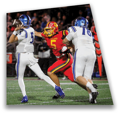 A person in football gear and helmet is crouched on a field with other players lined up in the background during a game. Description generated by AI