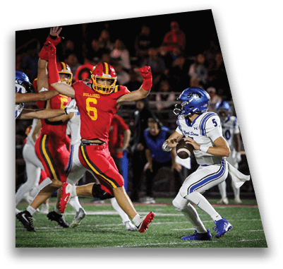 A person in football gear and helmet is crouched on a field with other players lined up in the background during a game. Description generated by AI
