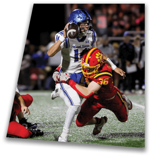 A person in a black football uniform and helmet is crouched in a ready position on a green field with another player in white uniform visible in the background. Description generated by AI