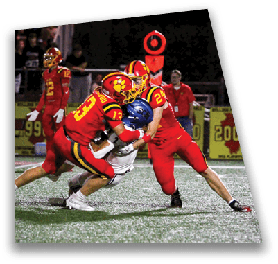 A person in football gear and helmet is crouched on a field with other players lined up in the background during a game. Description generated by AI