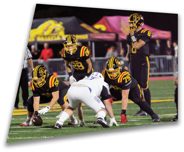 Four people in dark football uniforms and helmets stand in a row on a field, appearing focused and ready for action. Description generated by AI