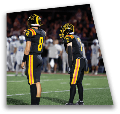 A person in football gear and helmet is crouched on a field with other players lined up in the background during a game. Description generated by AI