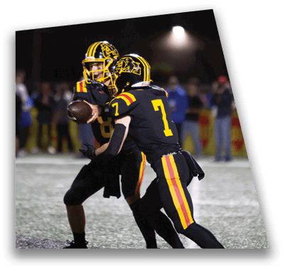 A person in football gear and helmet is crouched on a field with other players lined up in the background during a game. Description generated by AI
