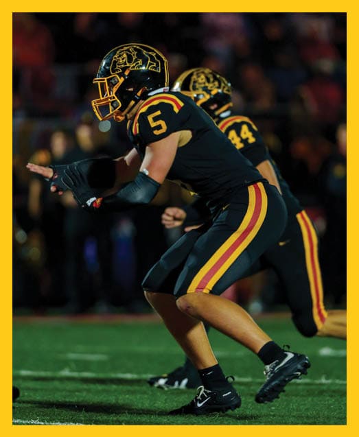 A person with curly hair wearing a black football uniform with “VISTA" on the jersey is sitting on a grassy field with one leg bent and the other extended. Description generated by AI