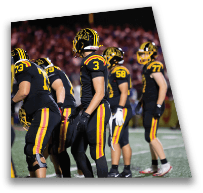 A person in football gear and helmet is crouched on a field with other players lined up in the background during a game. Description generated by AI