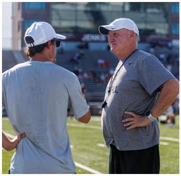 Two men wearing white hats and gray shirts are talking on a field. Description generated by AI