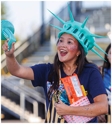 A woman wearing a blue shirt and a Statue of Liberty hat holds a blue and white frisbee. Description generated by AI