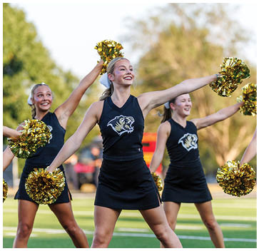 A cheerleader in a black and gold uniform is holding a yellow and black pom pom. Description generated by AI