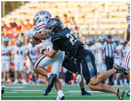 A football player in a black jersey is tackled by another player. Description generated by AI
