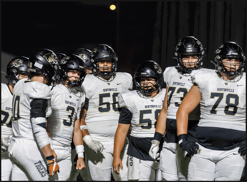 Four people wearing black football uniforms with “VISTA" on the front are standing together in a stadium with a crowd in the background, two of them holding helmets raised above their heads. Description generated by AI