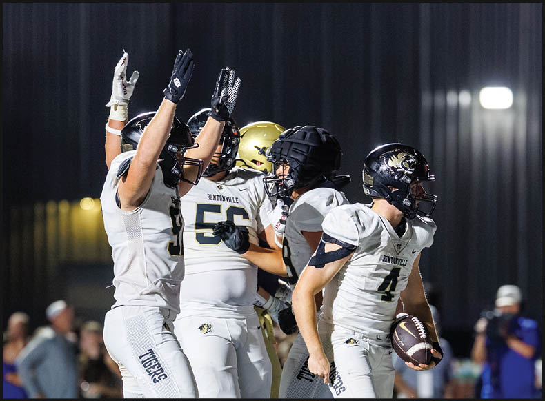 Four people wearing black football uniforms with “VISTA" on the front are standing together in a stadium with a crowd in the background, two of them holding helmets raised above their heads. Description generated by AI