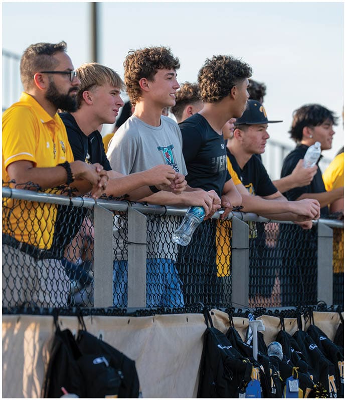 A group of people wearing casual and yellow sports attire are standing behind a chain-link fence, some holding water bottles, with backpacks hanging below the fence. Descripci n generada con IA