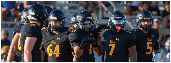 A group of five people wearing black football uniforms and helmets are standing together on a field with a crowd in the background. Descripci n generada con IA