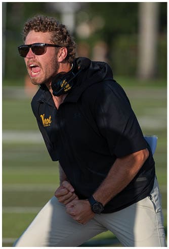 An adult wearing sunglasses and a black hoodie with a headset around their neck is shouting and clenching their fists on a sports field. Descripci n generada con IA