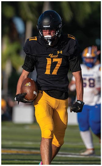 A person wearing a black football jersey with the number 17 and yellow pants is running on a field while holding a football. Descripci n generada con IA