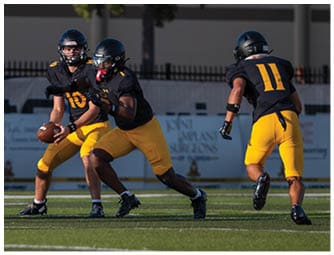 Three people wearing black helmets and black and yellow football uniforms are on a football field, with one person holding a football and another preparing to receive it. Descripci n generada con IA