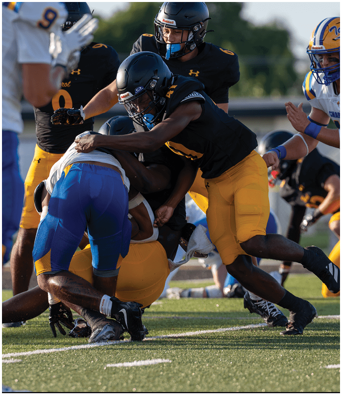 Several people wearing football uniforms are engaged in a tackle on a grassy field during a game. Descripci n generada con IA