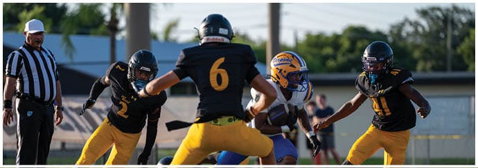 Four people in football uniforms are actively engaged in a play on a grassy field with one person holding the ball and others attempting to block or tackle. Descripci n generada con IA