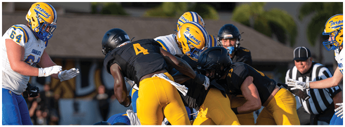 Several people in football uniforms are engaged in a close play on a grassy field with a referee observing nearby. Descripci n generada con IA