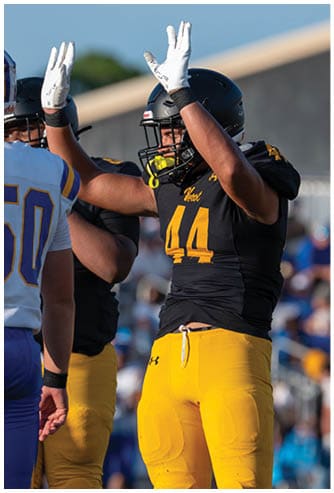 A person wearing a black football helmet and jersey with yellow lettering raises both arms while wearing white gloves during a game. Descripci n generada con IA