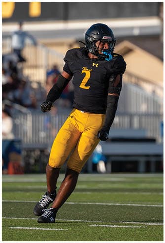 A person wearing a black football jersey with the number 2 and yellow pants is running on a football field during a game. Descripci n generada con IA
