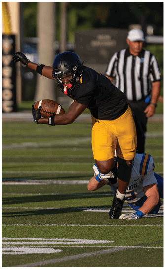 A person in black and yellow sportswear is running with a football while another person in blue and white sportswear is on the ground behind them and a referee stands in the background on a football field. Descripci n generada con IA